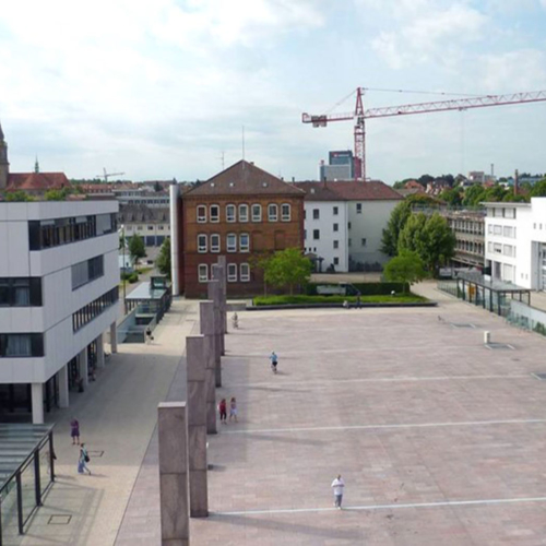 Das Foto zeigt den neuen Rathausplatz nach dem Umbau. Abriss und Neubau von Gebäuden sind zu vermerken. Auf dem Platz befinden sich sechs Betonstelen. Unter dem Platz ist eine Tiefgarage zu finden.